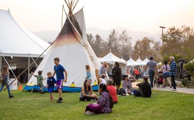 People of various ages sit and talk on the grass near a large tipi, with children playing nearby and several white tents in the background on a sunny day.