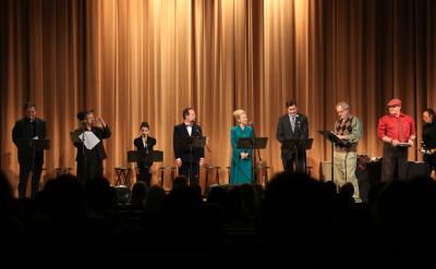 A group of people stand on stage in front of a gold curtain, each at a podium with scripts, performing a reading to an audience seated in the dark. The stage is brightly lit and the performers are dressed formally.