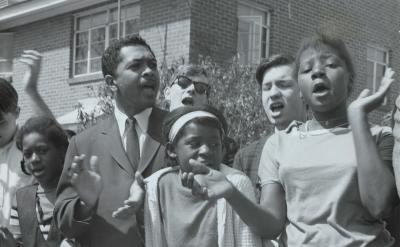A group of young people and a man in a suit stand together outside, clapping and singing with expressive faces in front of a brick building, appearing to participate in a civil rights demonstration.