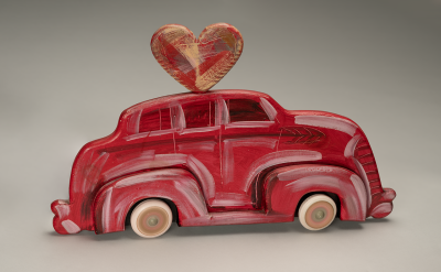 A toy car painted red with white accents, featuring a large red and gold heart shape attached to its roof, set against a plain gray background.
