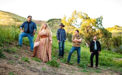 Five people stand on a grassy hillside, dressed in casual and country-style clothing. The woman in the center wears a patterned dress and boots, with trees and hills in the background under a clear sky.