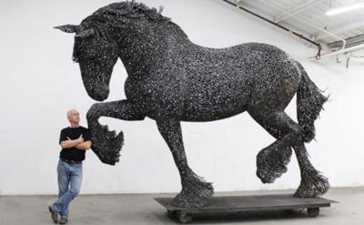 A man stands with arms crossed next to a large, detailed horse sculpture made from metal pieces, displayed indoors on a wheeled platform in a white-walled room with a high ceiling.