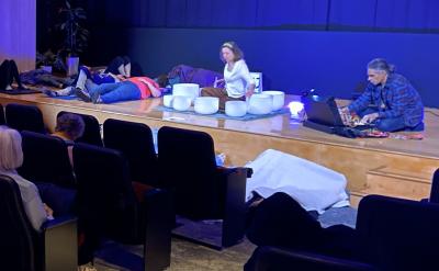 People sit in theater seats watching a sound bath performance on stage, where several individuals lie down, and two facilitators are playing crystal singing bowls under blue lighting.