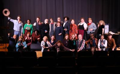 A group of around 25 people pose together on a stage, some standing and some sitting, in front of a black curtain. Several are smiling or gesturing enthusiastically, and the mood appears celebratory.