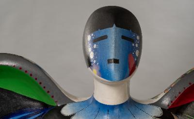 A close-up of a colorful abstract sculpture of a human figure with a smooth, painted face featuring blue and black geometric shapes, white dots, and bold green, red, and blue patterns on its shoulders.