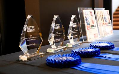 A row of glass awards labeled “Autry Museum” and 2023 sit on a black table alongside several blue ribbon rosettes, and framed certificates in the background.