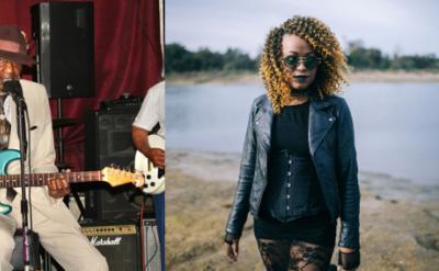 A man in a suit and hat plays an electric guitar and sings into a microphone on stage; beside him, a woman with curly hair wearing sunglasses, a black dress, and a leather jacket stands outdoors by water.