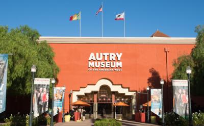 The entrance of the Autry Museum of the American West, with orange walls, three flags flying above, banners on either side, and people near the entrance on a sunny day.