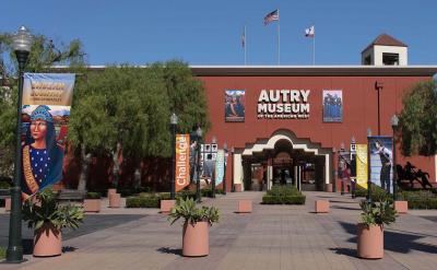 Entrance to the Autry Museum of the American West with banners reading "Indian Country," "Challenge," and "Discover," potted plants, and two flags flying above the red brick building.