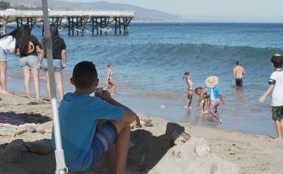 Children and adults enjoy a sunny day at the beach near a pier; some play in the sand, others wade in the water, and a boy in a blue shirt sits under an umbrella, watching the scene. The ocean and hills are in the background.