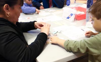An adult and a child sit at a table working on a craft project with paper, scissors, and other materials. Other children and adults are visible in the background, also participating in the activity.