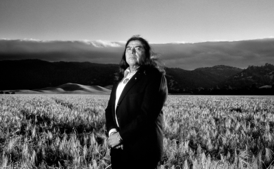 A man wearing a suit stands in a vast field of tall grass, with rolling hills and a cloudy sky in the background, captured in black and white.