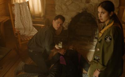 A young man kneels beside an unconscious or injured person lying on the floor of a rustic cabin, while a woman in a scout uniform stands nearby looking concerned. The room is warmly lit with wooden walls and furniture.