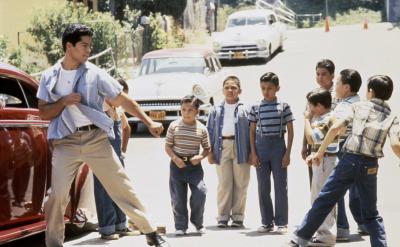A young man poses in a fighting stance near a red car on a street, facing a group of boys who watch him. The boys appear curious or excited, and old cars are parked in the background.