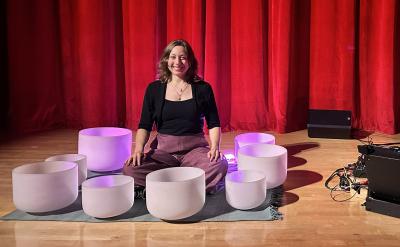 Two people sit on stage in front of a red curtain. One is surrounded by white crystal singing bowls, and the other sits beside electronic equipment. Both are smiling, seated on the floor under stage lighting.