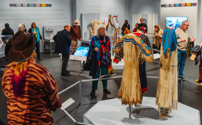 A group of people view traditional Indigenous clothing and artifacts displayed in a well-lit museum exhibit, with informational plaques and showcases on the walls and vibrant patterns featured on the garments.