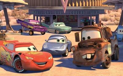 Animated cars with expressive faces gather at a gas station in a desert town. The group includes a red race car, a rusty tow truck, a blue car, a green van, and more, all smiling and interacting in front of a building.