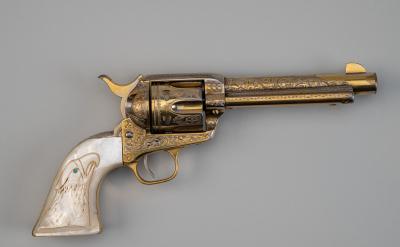 A decorative revolver with an engraved gold-colored barrel and cylinder, and a white handle featuring an eagle design and a blue gemstone eye, displayed on a plain gray background.