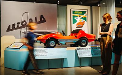 A red Disney Autopia car is displayed in a museum exhibit, with large Autopia signs and posters in the background. Two people stand nearby while another person walks past, slightly blurred.