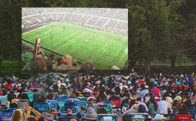 A large crowd sits outdoors on lawn chairs facing trees, watching a projected image of a stadium with a soccer game. The scene suggests a public outdoor viewing event.