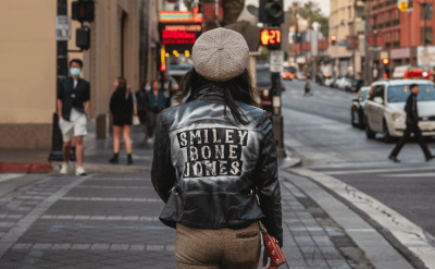 A person wearing a leather jacket with “SMILEY BONE JONES” on the back stands at a city street corner, facing away from the camera. Cars and pedestrians are visible in the urban background.
