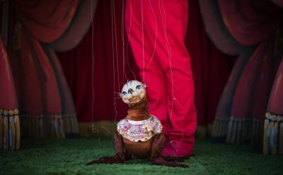 A marionette puppet dressed in a frilly outfit is controlled by strings, standing on green grass with a person in red pants behind it, in front of a stage with pink curtains.