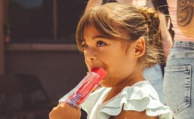 A young girl with brown hair in a bun enjoys a red popsicle outdoors. Adults stand nearby, one with a tattooed arm and another with keys clipped to their jeans. It appears to be a sunny day.
