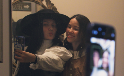 Two women pose for a selfie in front of a mirror; one wears a black hat and white blouse, and the other smiles in a tan vest. A phone held up in the foreground captures their reflection.