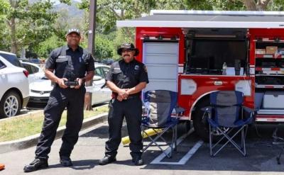 Two uniformed firefighters stand beside a red Los Angeles Fire Department command vehicle with open compartments displaying equipment. Folding chairs and traffic cones are set up nearby in a sunny outdoor area.