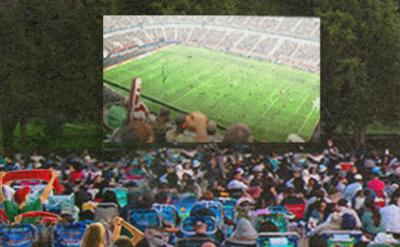 A large outdoor crowd sits on lawn chairs facing a screen showing a soccer match in a stadium. Trees line the background, creating a park-like setting.