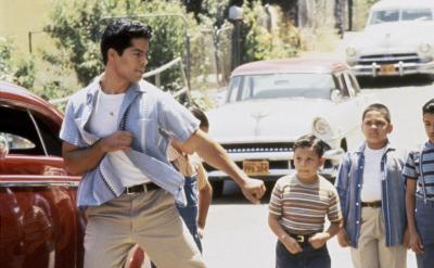 A teenage boy strikes a dramatic pose in front of a group of young boys on a sunlit street with vintage cars parked nearby. The boys watch him with curious and surprised expressions.