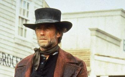 A man in a dark coat and tall black hat stands in front of old wooden buildings, including one marked “BANK,” with a western town setting and a serious expression on his face.