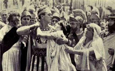 A group of people in ancient Roman attire surrounds a central figure being attacked, while others look on in shock or distress. The scene appears dramatic and intense, set on a crowded street.