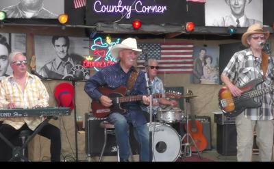 Four men perform country music onstage with guitars, keyboard, and drums. They wear casual clothes and hats. Neon signs, American flags, and black-and-white portraits decorate the background.