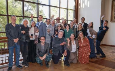 A group of people pose indoors near large windows, some standing and some kneeling, many smiling and waving at the camera. Sunlight streams in, and the setting has tiled floors and trees visible outside.
