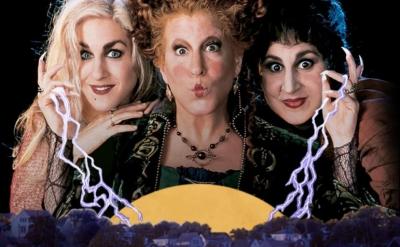 Three women in witch costumes make mischievous faces, holding glowing lightning above a town at night with a large yellow moon behind them.