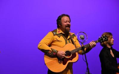 A man in a yellow shirt plays an acoustic guitar and sings into a microphone on stage with a purple background. Another musician stands beside him.