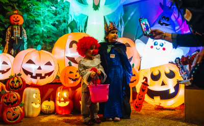 Two children in Halloween costumes stand among large glowing jack-o’-lantern decorations while someone takes their photo. The scene is colorful and festive with spooky, playful decorations.
