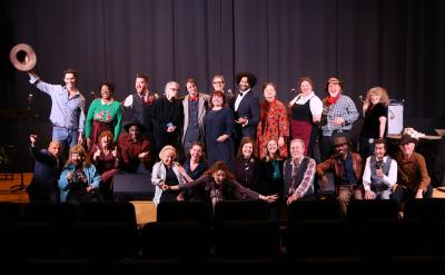 A group of about 25 adults pose and smile on a stage, some standing and some sitting, in front of a curtain. One person holds a hat in the air. The atmosphere is lively and cheerful.