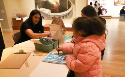 A young girl in a pink coat draws with colored pencils at a table, guided by an adult woman, in a brightly lit art gallery or museum space. Other children are also engaged in the activity.
