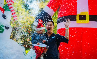 A man holding a toddler in a Santa hat while artificial snow falls in front of giant inflatable holiday decorations.