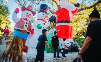 Three children stand in front of large inflatable Christmas decorations, including a cowboy snowman and Santa Claus, while artificial snow falls on them.