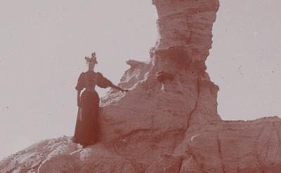 woman in 19th century clothing stands on steep hill next to rock formation