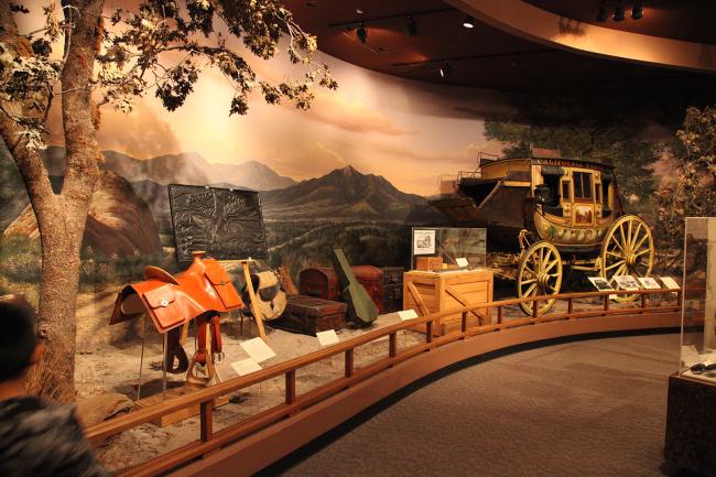 museum display of saddle and stagecoach with painted landscape background and a wooden rail in front