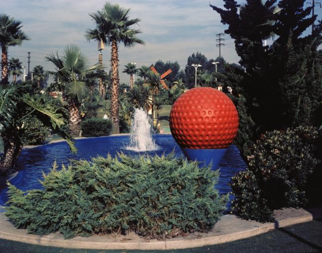 fountain in large pool of water with giant red golf ball on a giant blue tee n foreground