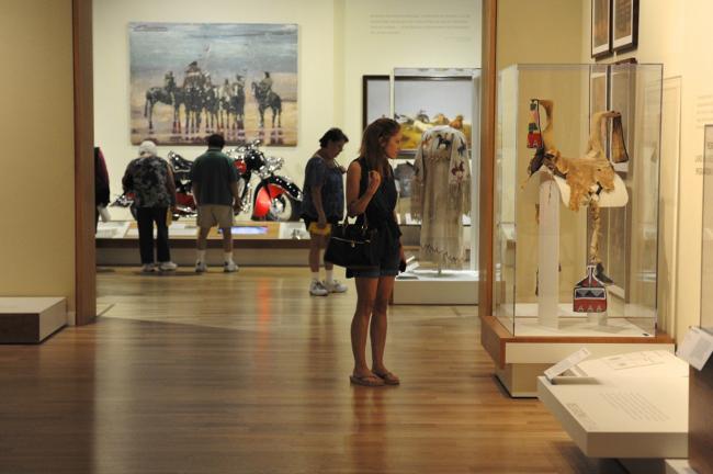a woman looking at a saddle on display in an exhibit with other people in the background looking at motorcycle on display in front of a painting of native americans on horseback