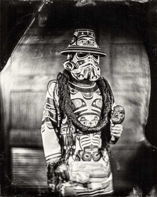 A person is dressed in a Star Wars stormtrooper helmet painted in Indigenous art style, wearing a matching robe, and holding a small carved mask. The background is blurred and the image has a vintage, black and white aesthetic.