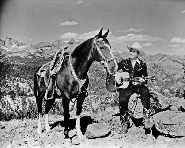 A man in cowboy attire sits on a rock in a mountainous landscape, playing an acoustic guitar. A saddled horse stands next to him. The sky is clear with scattered clouds, and the rugged terrain stretches out in the background.