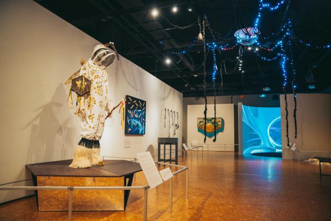 A dimly lit art gallery features a variety of exhibits. A centerpiece displays a mannequin adorned with an intricate, decorative costume. Art pieces hang on the walls. Blue lights and abstract decorations suspend from the ceiling, creating a mystical ambiance.