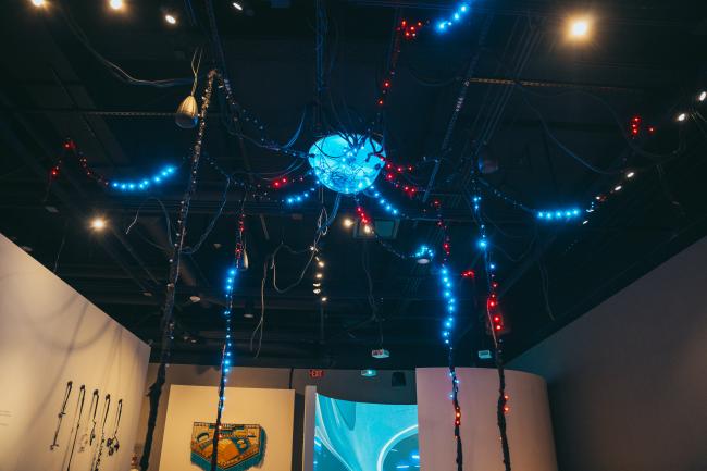 A ceiling installation resembling a spider with lighted red and blue cables extending from a central hub in a dimly lit room. The floor and surrounding walls feature various artworks and displays. The atmosphere is futuristic and somewhat industrial.